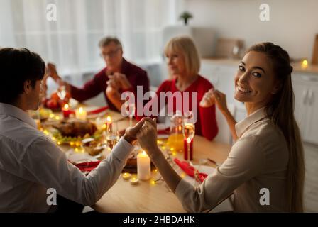 Young woman celebrating Christmas with her husband and senior parents, having festive Xmas dinner, praying to God Stock Photo
