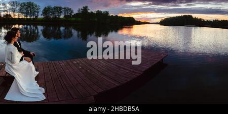 Wide angle panoramic view caucasian wedding couple sit together on bench enjoy lake and dramatic sunset panorama outdoors in Lithuania Stock Photo