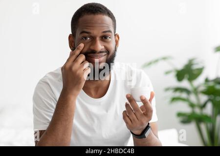 Happy young black bearded man hold jar and applying cream on face in white bedroom interior, close up Stock Photo