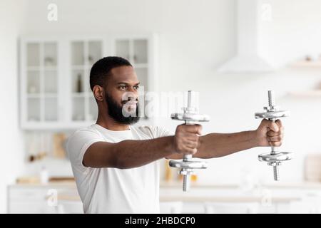Smiling young black bearded athlete man in white sportswear doing hands exercise with dumbbells Stock Photo
