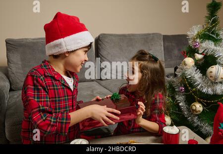 Adorable Caucasian children, cute boy in Santa's hat and charming baby girl sit at table with freshly baked Stollen bread and hot drink with marshmall Stock Photo