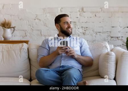 Happy dreamy young man using smartphone at home. Stock Photo
