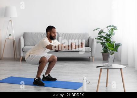 Concentrated young black bearded athlete man in white sports clothes squatting in living room interior, profile Stock Photo