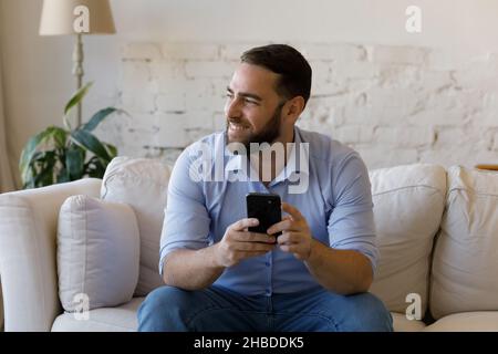 Smiling relaxed dreamy young man using smartphone at home. Stock Photo