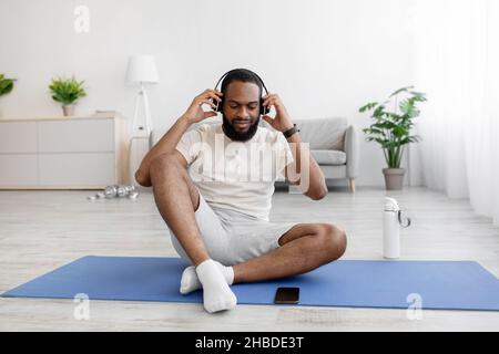 Smiling young black bearded athlete man in white clothes sits on mat with bottle of water Stock Photo