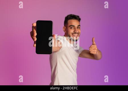 Cheerful millennial Arab guy pointing at smartphone with blank screen, recommending mobile app in neon light, mockup Stock Photo