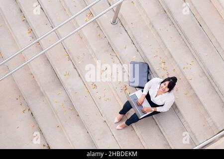 top view of a smiling business woman typing on her laptop computer sitting on a concrete staircase, concept of digital entrepreneur and urban lifestyl Stock Photo