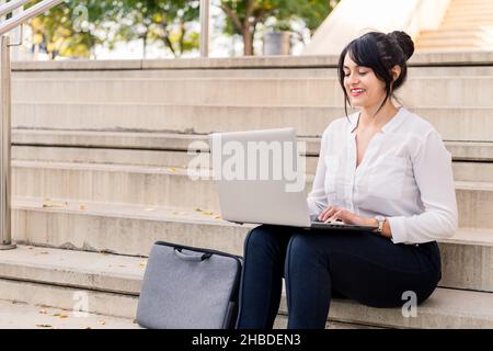 smiling businesswoman working on her laptop computer sitting on a concrete staircase, concept of digital entrepreneur and urban lifestyle, copy space Stock Photo
