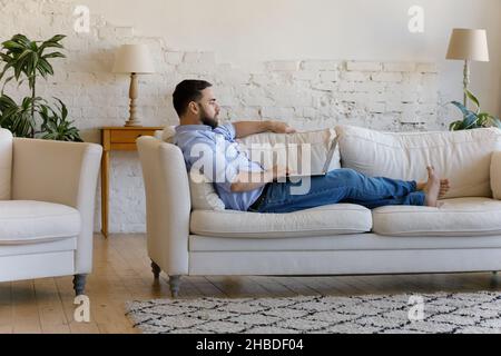 Concentrated young man using computer lying on couch. Stock Photo