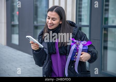 Young woman with a purple gift in hands uses a smartphone outside. Stock Photo