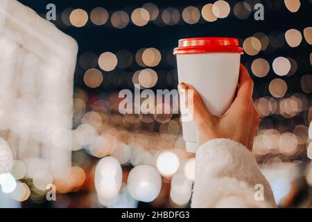 Female hands holding a plastic cup with hot tea or coffee against the background of bokeh garland lights. Christmas holiday magic details Stock Photo