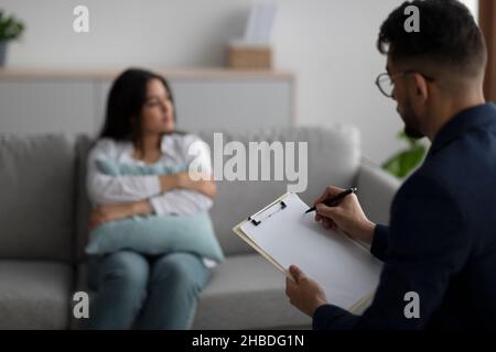 Sad young arab woman hugging pillow and talking to psychologist, focus on psychiatrist taking notes in clipboard Stock Photo