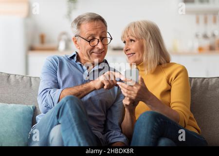 Senior Couple Texting Via Smartphone Sitting On Sofa At Home Stock Photo