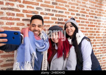 Positive multiracial male and female friends in stylish outfits smiling brightly while taking self portrait on cellphone near brick cladding Stock Photo