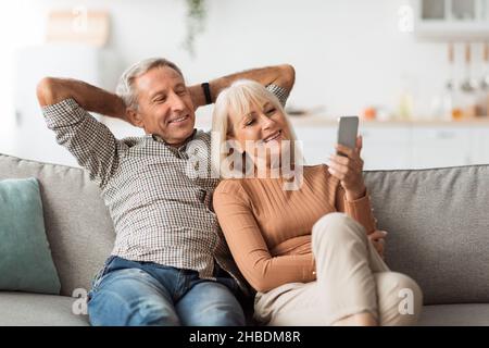 Happy Senior Spouses Using Mobile Phone Sitting On Couch Indoor Stock Photo