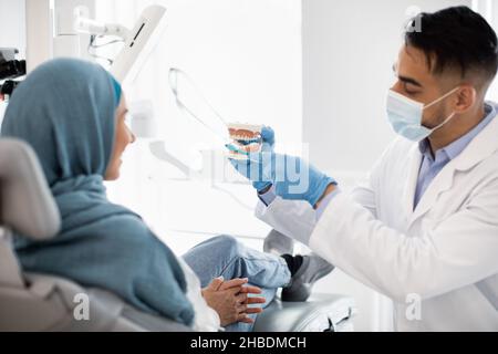 Arab Stomatologist Holding Jaw Model, Teaching Oral Hygiene To Muslim Female Patient Stock Photo