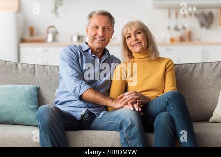 Older Spouses Holding Hands Sitting On Sofa Posing At Home Stock Photo