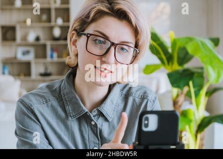Woman blogger with glasses and a gray shirt talks in front of the phone an online lecture in a cozy office with houseplants Stock Photo