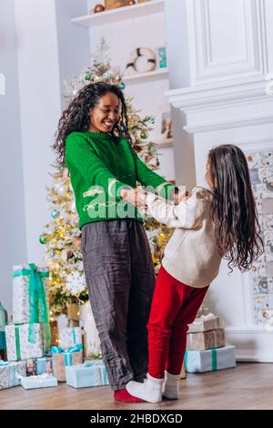 Brunette long-haired daughter plays and dances with African American mother smiling happily near Christmas tree with gift boxes Stock Photo