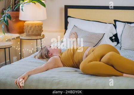 Woman with long blonde hair wearing matching sports set lies on bed with blanket and pillows relaxing after yoga workout in bedroom Stock Photo