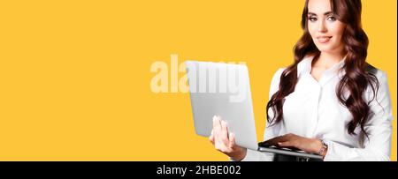 Beautiful positive girl with laptop in her hands on yellow background looks at camera and smiles. Caucasian woman with loose hair of 25-30 years old in white blouse and open laptop. Advertising banner with space for copying. Stock Photo