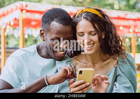 Young Afro-American man and brunette young woman watch selfy on smartphone in attraction park on sunny summer day closeup Stock Photo