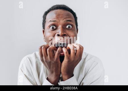 Portrait of a terrified African man staring in horror at the camera and putting his hands to his mouth in a light linen shirt on a white background. Very strong surprise or fright, horror in the eyes. Stock Photo