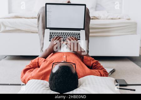 African-American man blogger in orange shirt and glasses works on laptop with blank screen lying on floor at home close upper back view Stock Photo