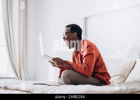 Emotional positive black man in colorful clothes and glasses screams holding modern laptop on large bed at home side view Stock Photo