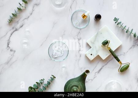 Handmade moisturizer and green jade face roller. Cotton towel. Exotic eucalyptus leaves and water drops on off white marble background with stone Stock Photo