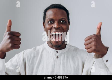 Portrait of emotional happy African-American guy in white shirt showing thumbs up posing on light grey background close view Stock Photo
