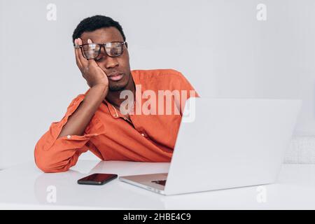 African man in an orange shirt and glasses sleeps leaning on his arm sitting at his desk in front of a laptop and phone on a light background. Young freelancer tired of working on a project, a break in work Stock Photo