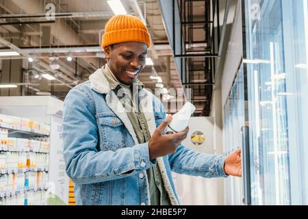 Satisfied African millennial man in an orange hat in a supermarket chooses bottled milk standing at the refrigerator, and reads the composition of the product, smiles found the right product Stock Photo