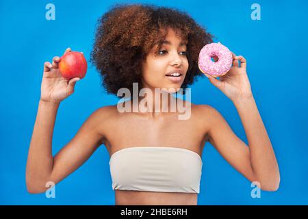 Curly haired mixed race young woman wearing light top poses holding apple and donut with frosting and smiles against bright blue wall Stock Photo