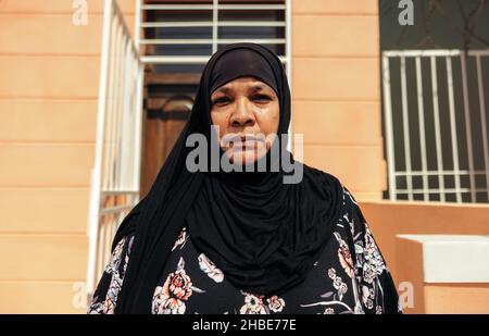Middle-aged Muslim woman looking at the camera confidently while standing in front of her home during the day. Self-confident mid-adult woman wearing Stock Photo