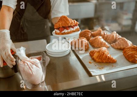 Confectioner holds croissant with juicy strawberry at metal counter in bakery Stock Photo