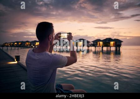 Young man with mobile phone photographing seascape with water bungalows at beautiful sunset in Maldives. Stock Photo