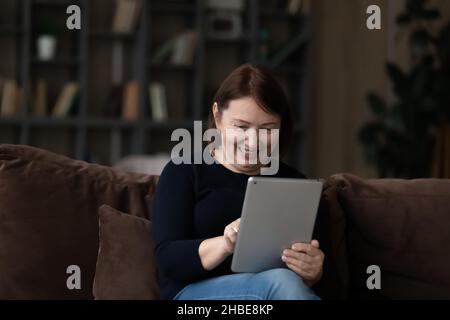 Aged female pensioner sit on sofa hold touchpad surf internet Stock Photo