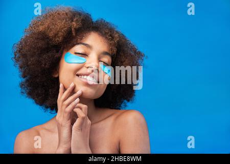 Curly-haired brunette young woman smiles widely with bright blue beauty patches against pouches under eyes on background of studio wall Stock Photo