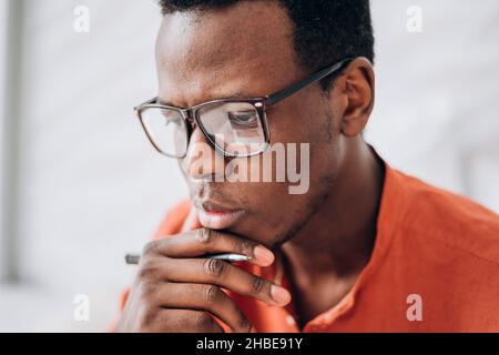 Thoughtful young African-American man with elegant glasses and pen wearing bright orange clothes in light room close view Stock Photo