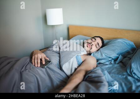 Young fit millenial multiracial man has bad dream in the morning with phone on stomach, stomach ache, having trouble sleeping in. 30-something guy Stock Photo