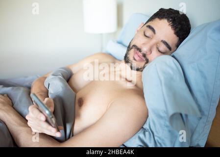 Mixed-race young 30-something millenial man laying in bed propped up on pillows looking at phone. Checking phone when waking up in the morning Stock Photo
