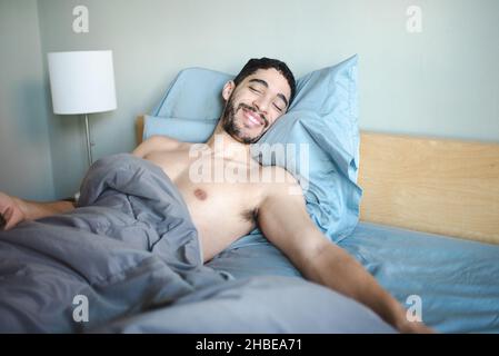 Mixed-race millenial bearded 30-something man smiles while propped up with two pillows in bed with light blue sheets and grey comforter, eyes closed Stock Photo