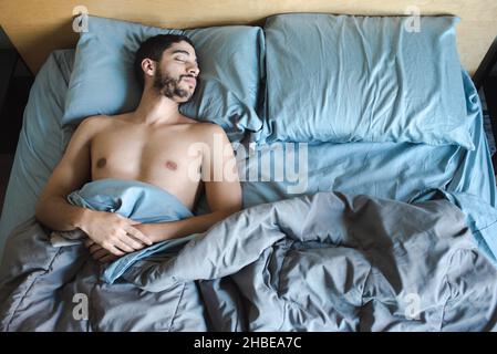 Multiracial young thin bearded millenial man sleeping on left side of the bed, empty space where girlfriend or partner was, in the mornign light Stock Photo