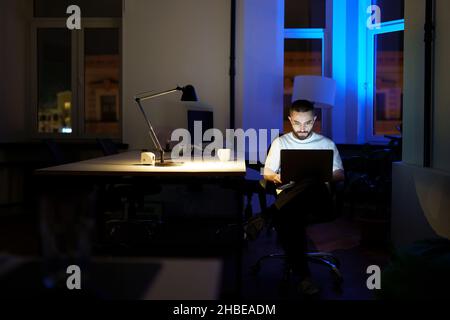 Sleepless programmer work on laptop at night. Young specialist in eyewear with nighttime lifestyle Stock Photo