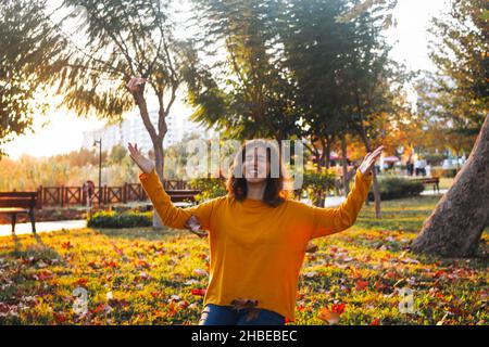 Curly young girl in yellow sweater and jeans sitting on grass and throws up dry leaves. Stock Photo