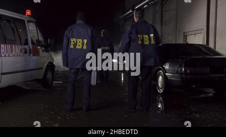 FBI agents work at the scene at night, police car with lights and ambulance background. Back view on three FBI agents go towards criminal scene. Stock Photo