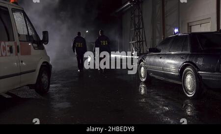 FBI agents work at the scene at night, police car with lights and ambulance background. Back view on three FBI agents go towards criminal scene. Stock Photo