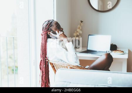 A woman sitting on a chair at the table and holding her smartphone. A young black female with braided hair wearing a white shirt, sitting and talking Stock Photo