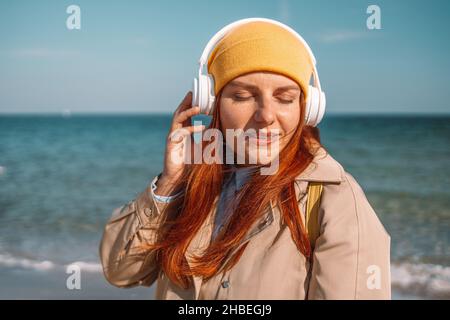 Lifestyle portrait of stylish Caucasian red hair woman in yellow cap wearing headphones listening music or podcast from smartphone application against Stock Photo